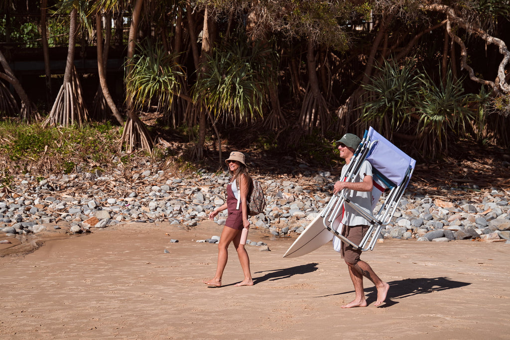 Two people walking on a beach with a surfboard and chair, surrounded by trees.