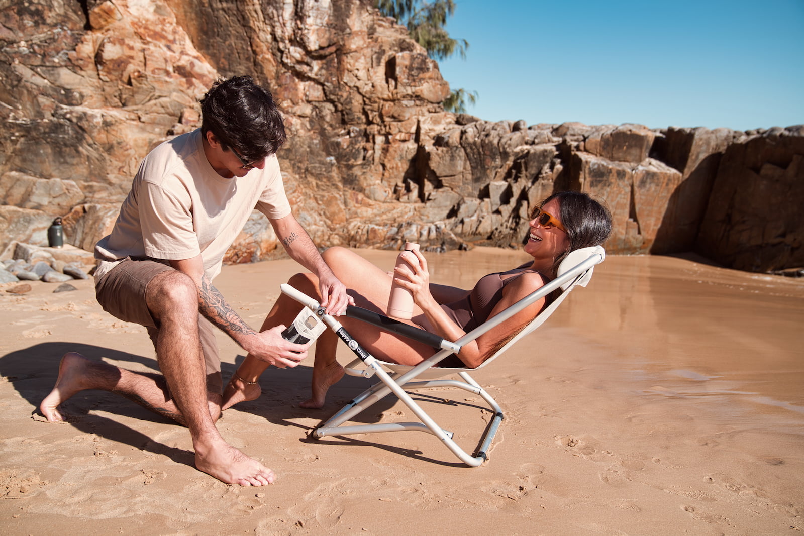 Two people by a rocky beach with a man attaching a cup holder to a woman's beach chair