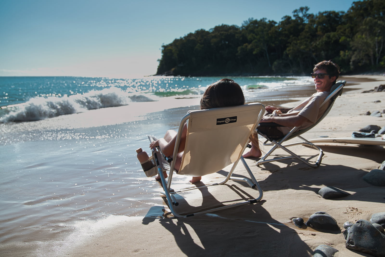 Two people sitting on happy chap beach chairs by the ocean with waves crashing onto the shore.