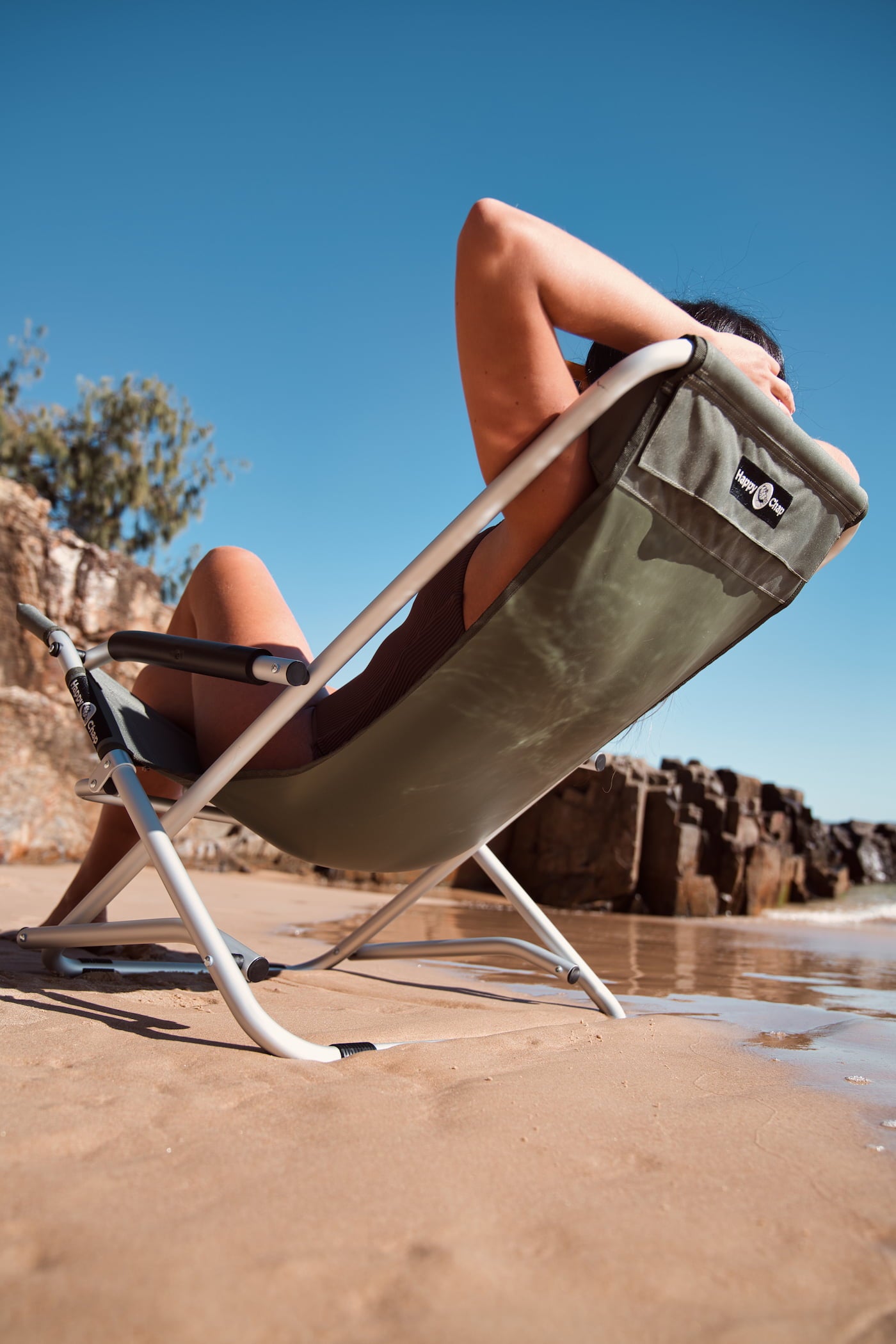 Person relaxing on a folding chair on a beach with a clear blue sky.