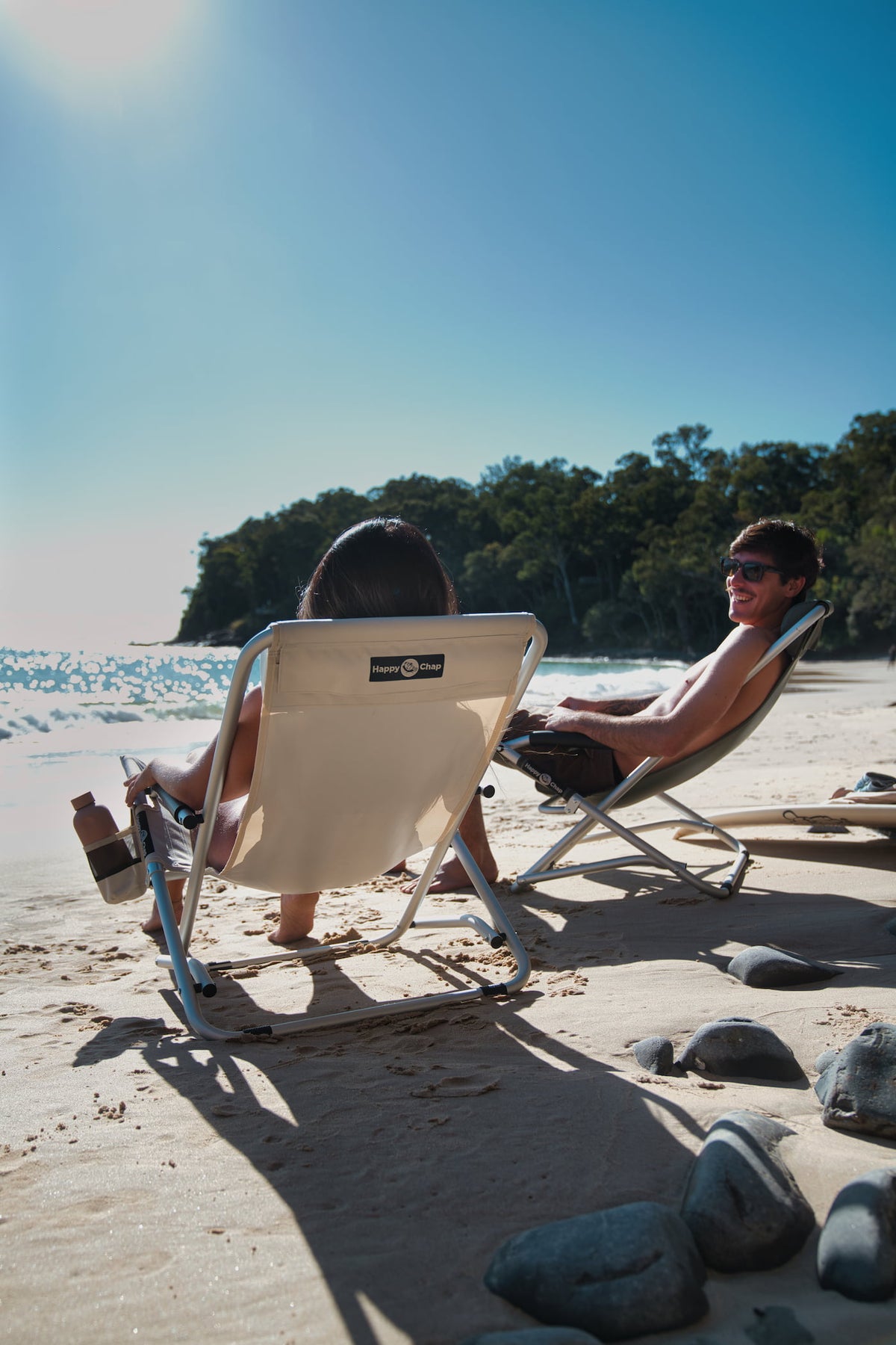 Two people sitting on comfortable beach chairs by the water with a clear blue sky.